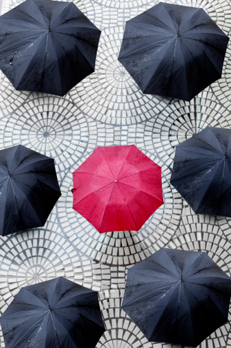 A single, solitary red umbrella surrounded by bleak, black umbrellas on a circular tile background.