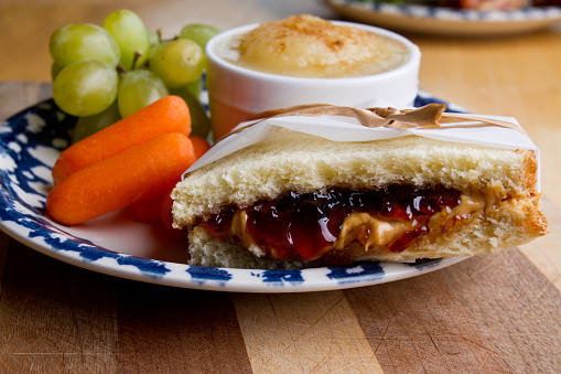 Classic kids lunch of peanut butter and jelly sandwich wrapped and tied elegantly in a paper wrapper.  Sandwich is on a retro blue plate on a cutting board with peeled baby carrots, green grapes and cinnamon topped apple sauce in a round white ramekin.