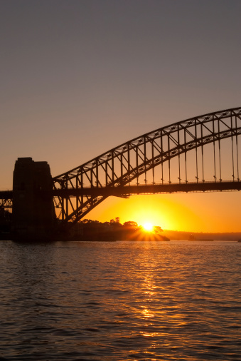 The sun appears on the horizon behind the Sydney Harbour Bridge at dawn.