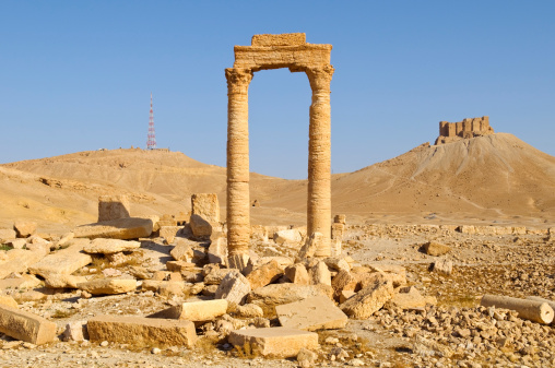 Ancient ruins and a modern communication tower in Palmyra, Syria. In the foreground are columns and rubble from the late Roman period or shortly thereafter; on the hill on the right is a 17th century fort; on the hill on the left is a modern communication tower.