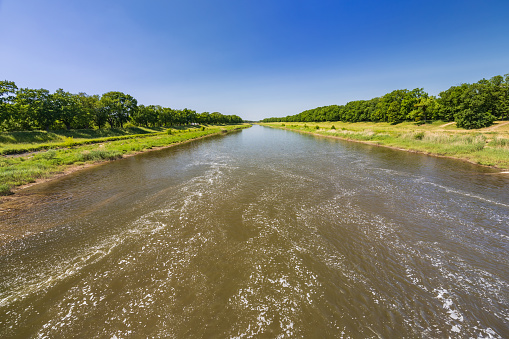 Beautiful landscape of long dirty river with green trees and bushes on both sides seen from bridge over water dam and water level