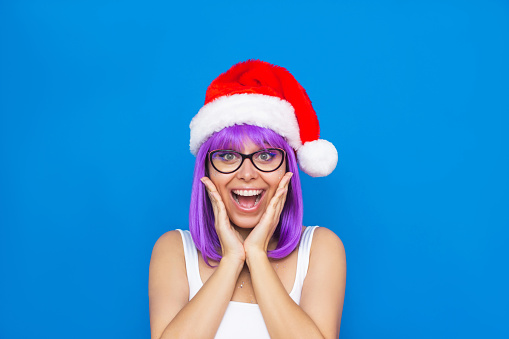 A young caucasian amazed smiling blonde woman with violet hair and red Santa Claus hat holding her hands to her face isolated on a color blue background. Christmas and New Year concept
