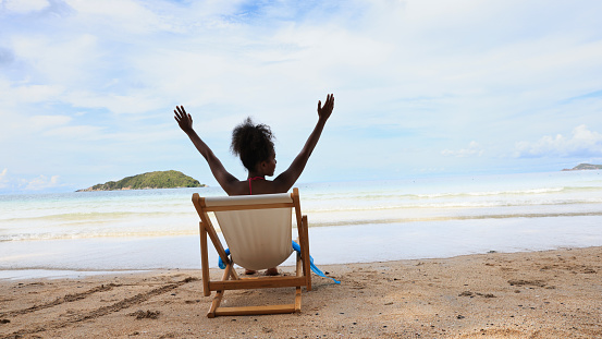 A Black woman is enjoying herself as she sits and relaxes on a white sun chair on a tropical beach.