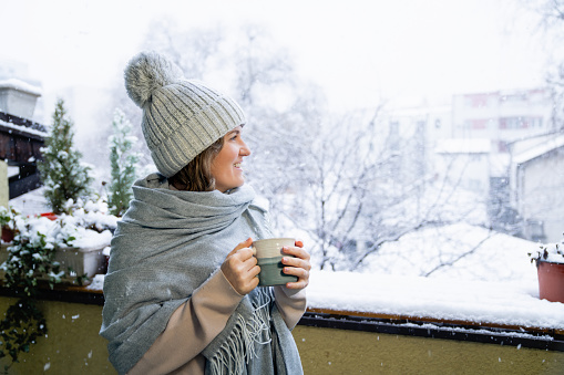 A woman in a warm hat and blanket holds a mug with a hot drink in her hands. Winter landscape with snowfall in the background.