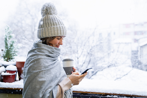 A woman in a warm hat and blanket holds smartphone in her hands. Winter landscape with snowfall in the background.