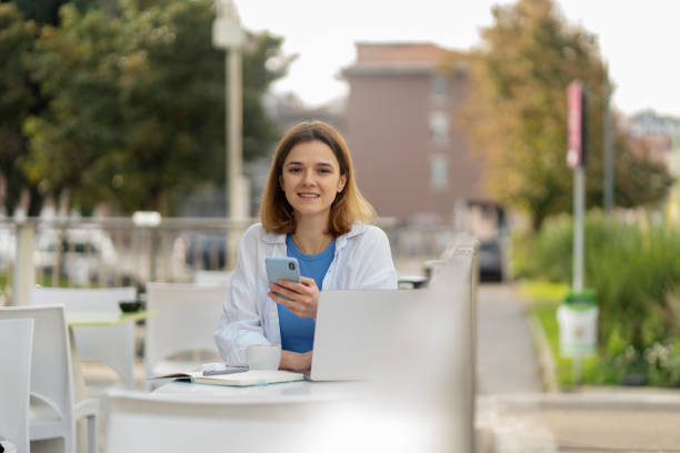 istock Smiling woman using mobile phone looking at camera sitting in cafe terrace 1711856578