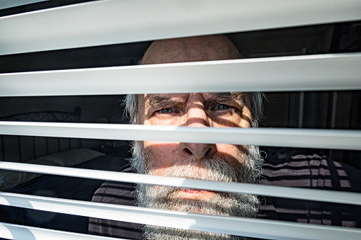 A suspicious senior adult man is staring warily - looking out through contrasting sun and shade stripe window blinds.