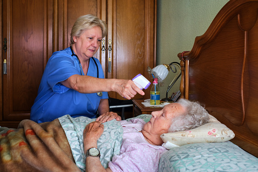 A mature nurse taking care of an older woman taking the body temperature in her bed.