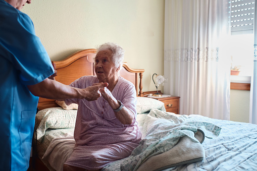 Side view of an unrecognizable nurse helping to stand up an elderly woman who is sitting on her bed in her bedroom at home with copy space.