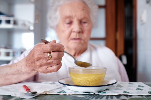 Front view of a old woman sitting in her kitchen eating a nutritious soup.