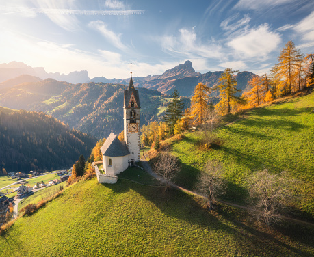 Aerial view of beautiful church, green alpine meadows, orange trees, hills in mountain village at sunset in autumn. Dolomites, Italy. Top view of old chapel, rocks, forest, sky with clouds in fall