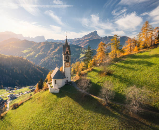 Aerial view of beautiful church, green alpine meadows, orange trees, hills in mountain village at sunset in autumn. Dolomites, Italy. Top view of old chapel, rocks, forest, sky with clouds in fall stock photo