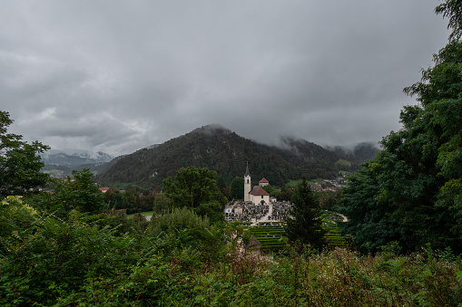 Cemetery and main church at the city of Kamnik, with green foliage in the foreground and castle hill in the far back. Cloudy murky setting.
