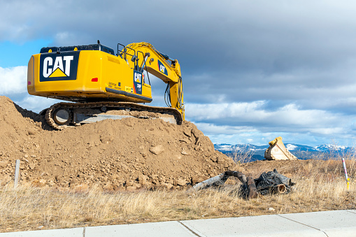 General view of a CAT 336F Sand hydraulic mining shovel on top of a mount of dirt on a hilltop with snow covered mountains in the distance, in Spokane, Washington.