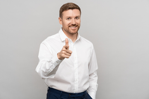 Joyful businessman with smile pointing index finger at camera isolated on studio gray background. Concept of hiring, employment and career success. Employer, headhunter, HR manager choosing you.