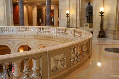 Interior upper floor circular balcony and hall walkway of the State Capitol building in St. Paul, Minnesota, USA. The legistlative building is at the heart of local politics and government. Marble columns, monument statues of historical figures, and the American flag give the built structure a sense of dignified importance.