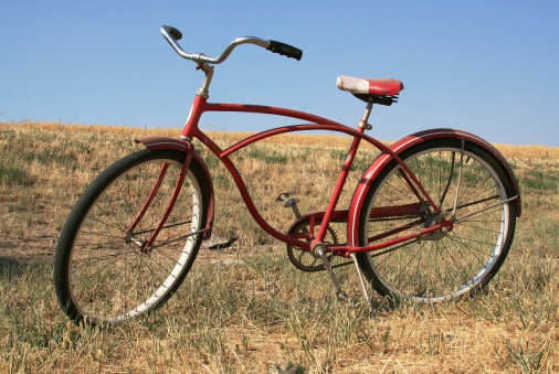 A vintage red bike rests on its kickstand with blue sky and grass behind it.