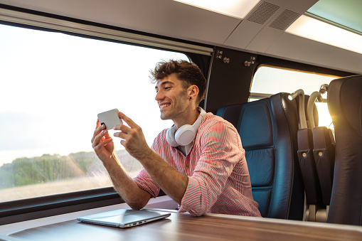 Portrait of a cheerful man in smart casual clothes watching something on smart phone while traveling by train. His laptop is on the table,