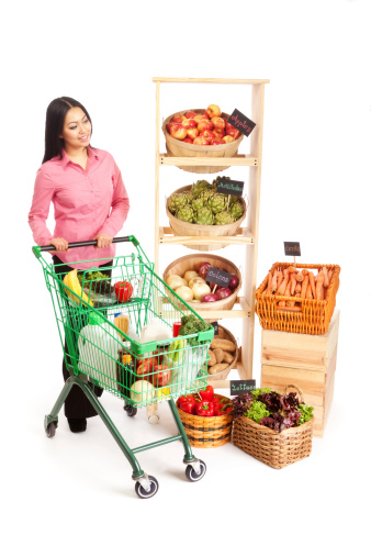 Subject: Happy smiling Asian woman pushing a grocery cart filled with fresh fruit, vegetables, and other store items. Isolated on white background