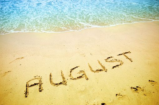 August written on the sand of a beautiful tropical beach. Visible are one island in the sea, turquoise water, little splashy waves, golden sand and beautiful cloudscape over the sea.