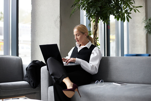 Attractive blonde businesswoman using laptop computer, typing on keyboard, working freelance project at workplace. Portrait of middle aged stylish secretary checking email sitting in modern office