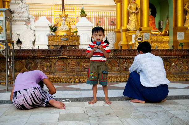Yangon, Myanmar - November 15, 2011: A Burmese boy plays with a cell phone as his parents pray at the landmark Shwedagon Pagoda in central Yangon. This pagoda is revered by Buddhist throughout Myanmar.