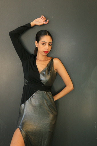 A Moroccan model dressed in a formal asymmetrical gown. She is wearing medium length black hair, makeup and a black and silver asymmetrical ball gown. There is a gray wall in the background.