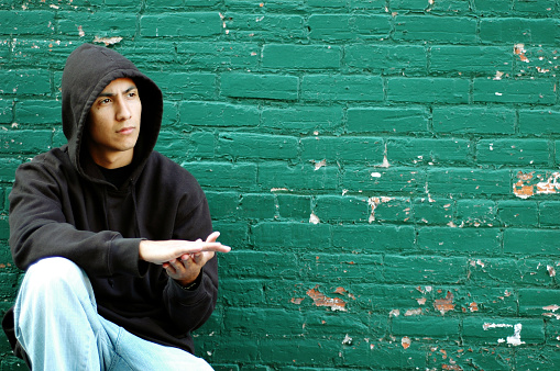Color photo of a young, hispanic man in an alley crouching by a green brick wall thinking about his life on the street as a gang member.