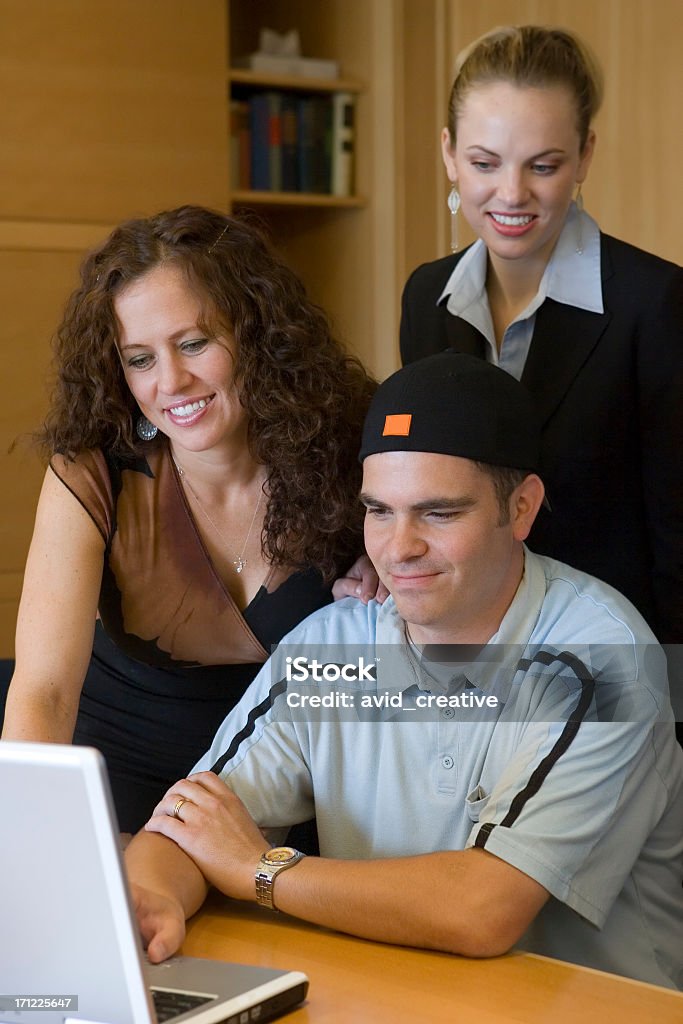 Happy Colleagues Three young business people collaborate on a project around a laptop in a boardroom. He could be collaborating with them, helping them prepare their presentation, showing them how to use the computer, or fixing a problem. Natural light. Shallow depth of field. Focus is primarily on the IT guy (young man in blue shirt) and the brunette. Problems Stock Photo