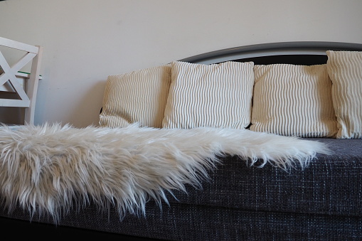 Gray sofa with boucle upholstery fabric and white decorative fluted cushions. White rug or bedspread made of faux fur with a long pile. Living room interior. Wooden painted chairs