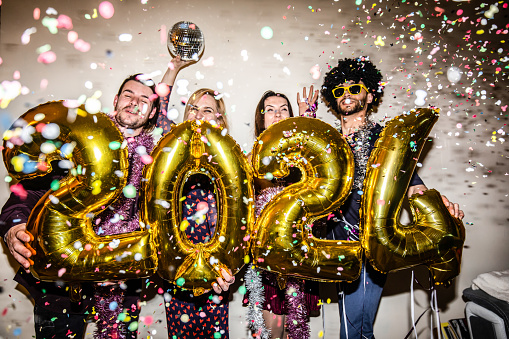 Four friends are celebrating the New Year 2024 with a party and big shiny foil balloons for 2024 and lots of falling confetti