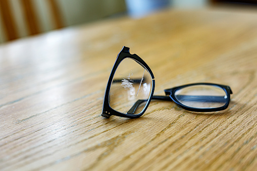 A pair of completely separated horn rimmed eyeglasses on an oak wood dining room table top. The glasses were broken in two when a senior man stumbled and fell while walking on a concrete footpath.