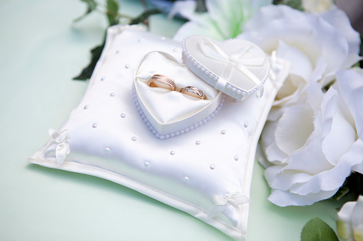 Close-up of white heart-shaped jewelry box with a wedding rings and white flowers in the background.