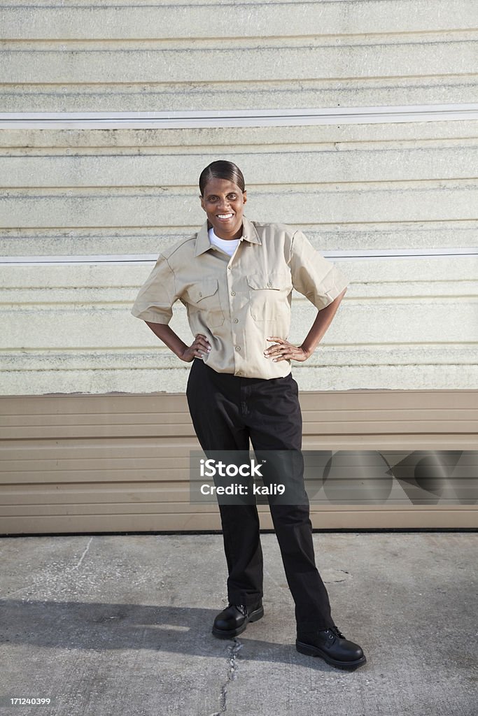 Female worker standing in front of warehouse door African American female worker (50s) standing in front of warehouse door. Full Length Stock Photo