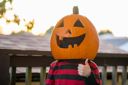 Anonymous child dressed in a red and black striped shirt is wearing a carved pumpkin jack-o-lantern on his head to celebrate the Halloween holiday in October outdoors. He is giving a thumbs up at the camera and the pumpkin is carved to be smiling a toothy smile.
