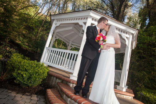 This is a vertical color image of newlyweds Kissing outside a gazebo.  There are some woods in the background