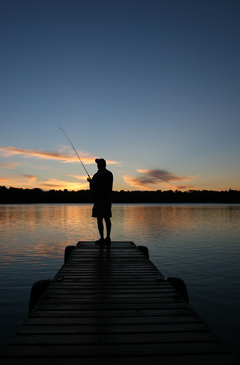 A retired senior man fishes off a dock at sunrise. Vertical colour image. Man is casting his lure into a pristine lake off the end of a wooded dock. Back view. Man is unrecognizable in the image. Themes include seniors, retirement, active living, lifestyle, dock, cottage, fishing, recreation, leisure, weekend, rod and reel, catching fish, and healthy living. 