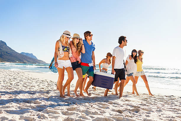Group of people carrying cooler to party on beach A group of seven young men and women walking down a beach.  The woman at the far left is has blond hair and is wearing a cream-colored hat with a black band.  The woman next to her is wearing a pink shirt.  The man in the middle is wearing a blue shirt and carrying one side of a cooler.  The man next to him is wearing a white shirt and holding the opposite side of the cooler.  The two women at the far end of the group have their arms around another.  A woman wearing a white top and brown hat walks slightly behind the group. beach party stock pictures, royalty-free photos & images