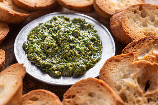 Homemade pesto sauce in a glass jar with basil, ingredients and baguette on white table in the kitchen. Pesto - traditional italian sauce for fettuccine, focaccia, pizza, toast, sandwiches