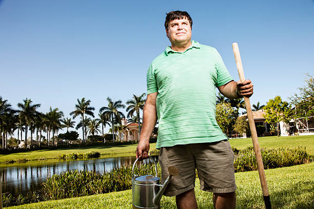 An experienced senior gardener in a straw hat, blue shirt, and jeans dungarees, casually leaning with work gloves on a spade against a white background  a symbol of dedicated gardening.
