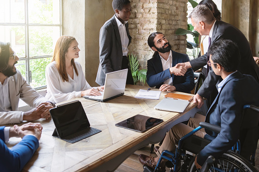 Young bearded man and senior silver-haired individual seal a deal amid diverse colleagues, including an inclusive member in a wheelchair. Bright office ambiance.