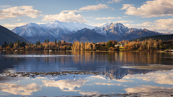 Looking across Lake Wanaka to the eastern shore of the Wanaka township, with snow-capped peaks in the background. The lake level is low, creating still pools of water that show perfect reflections of the mountains.