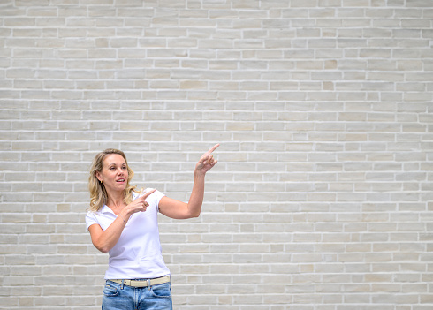 Promotional photo of an attractive friendly woman pointing to the right side and up in front of a white brick wall