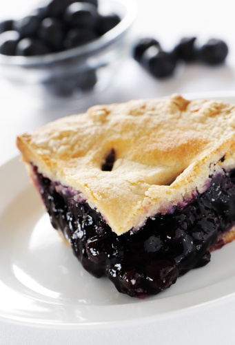Serving or slice of blueberry pie with blueberry out of focus in background.
