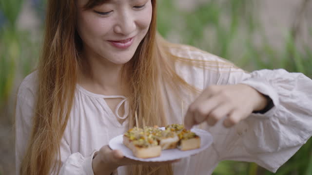 A beautiful woman holding a plate of canape', eating and feeling happy with the taste while showing the plate to the camera with a smile on her face during her lunch at a steakhouse with friends.