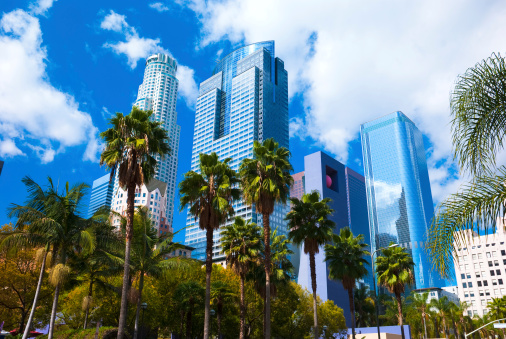 Los Angeles downtown skyscrapers with palm trees and Pershing Square in the foreground with clouds and a blue sky in the background.