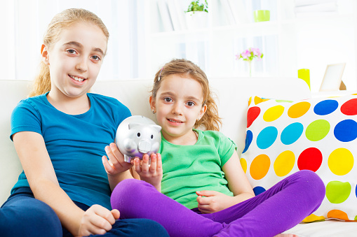 Portrait of two cute little girls, sisters, sitting on sofa and holding their savings piggy bank.