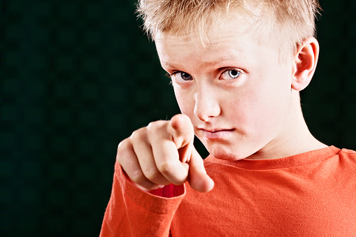 A good looking 9 year old blond boy with eyebrows raised points a finger at camera accusingly against a black background with copy space. 