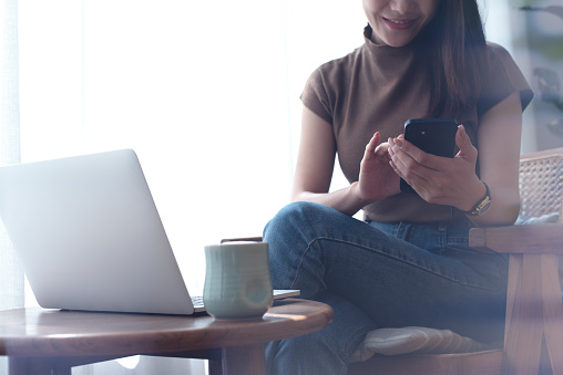 Smiling asian woman in casual wear using mobile phone while working remotely on laptop at coffee shop. Young woman freelancer online working and using smartphone, surfing the internet, freelance at work, vintage style