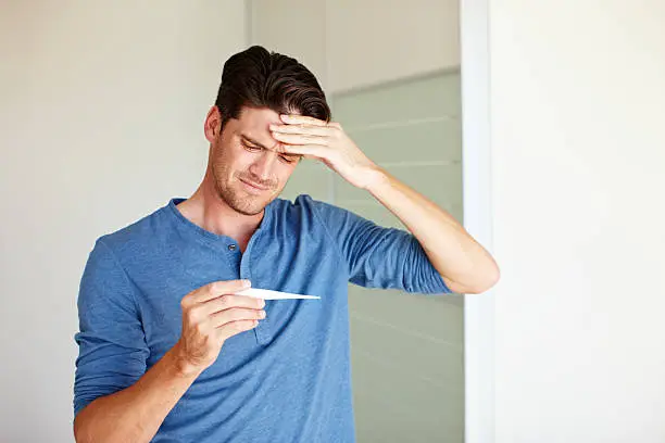 Unwell man holding his forehead while seeing that he has a fever on a thermometer